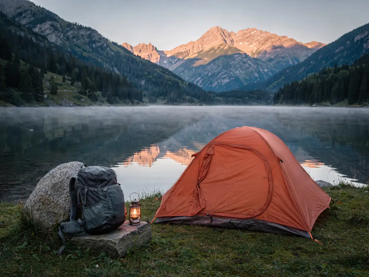 An orange tent pitched beside a calm mountain lake at dawn, with a backpack and a lantern beside it